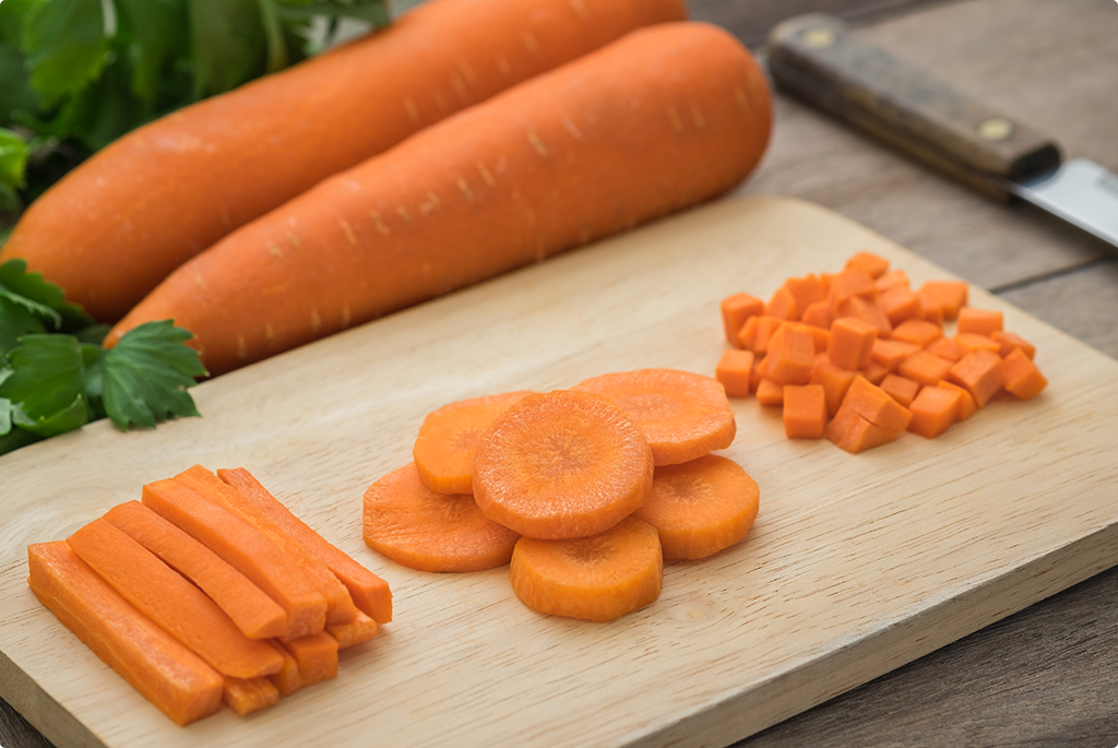 Two whole carrots, and a chopping board with one sliced into sticks, circles and diced on a wooden chopping board