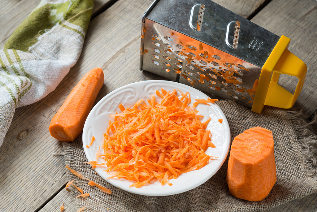 Grated carrot in a white bowl on a wooden table with grater and carrot next to it